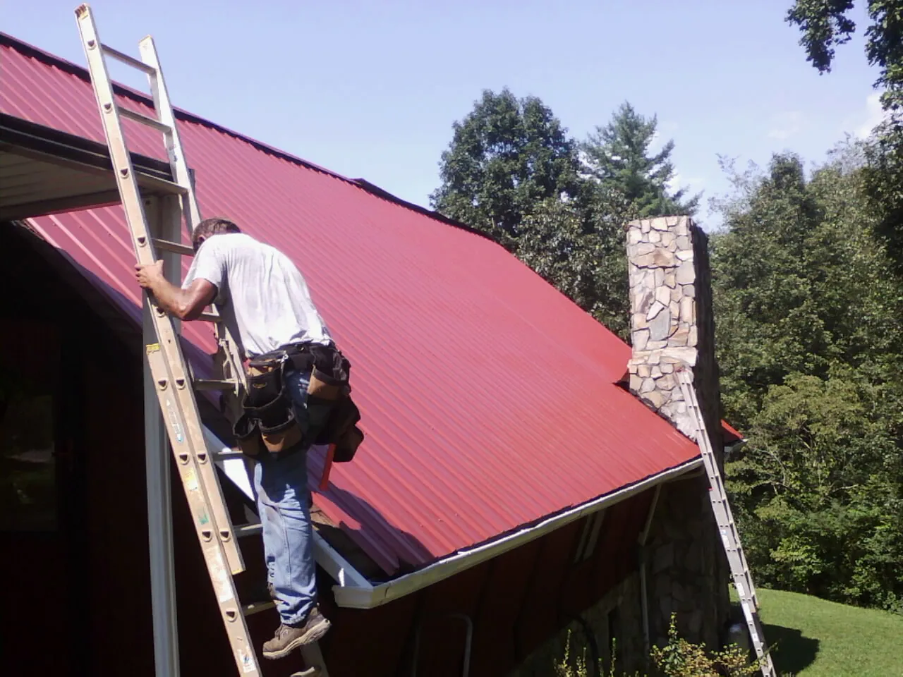 Roofer on a ladder beside a red metal roof and stone chimney — Soffit & Fascia Repair by Roofing Brothers in Swansboro